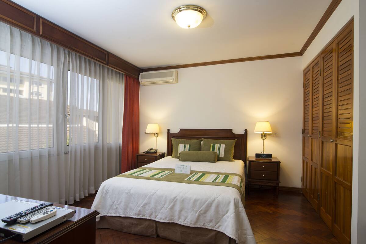 A hotel room with a bed, bedside lamps, a desk with a laptop and remote controls, and a window with curtains.