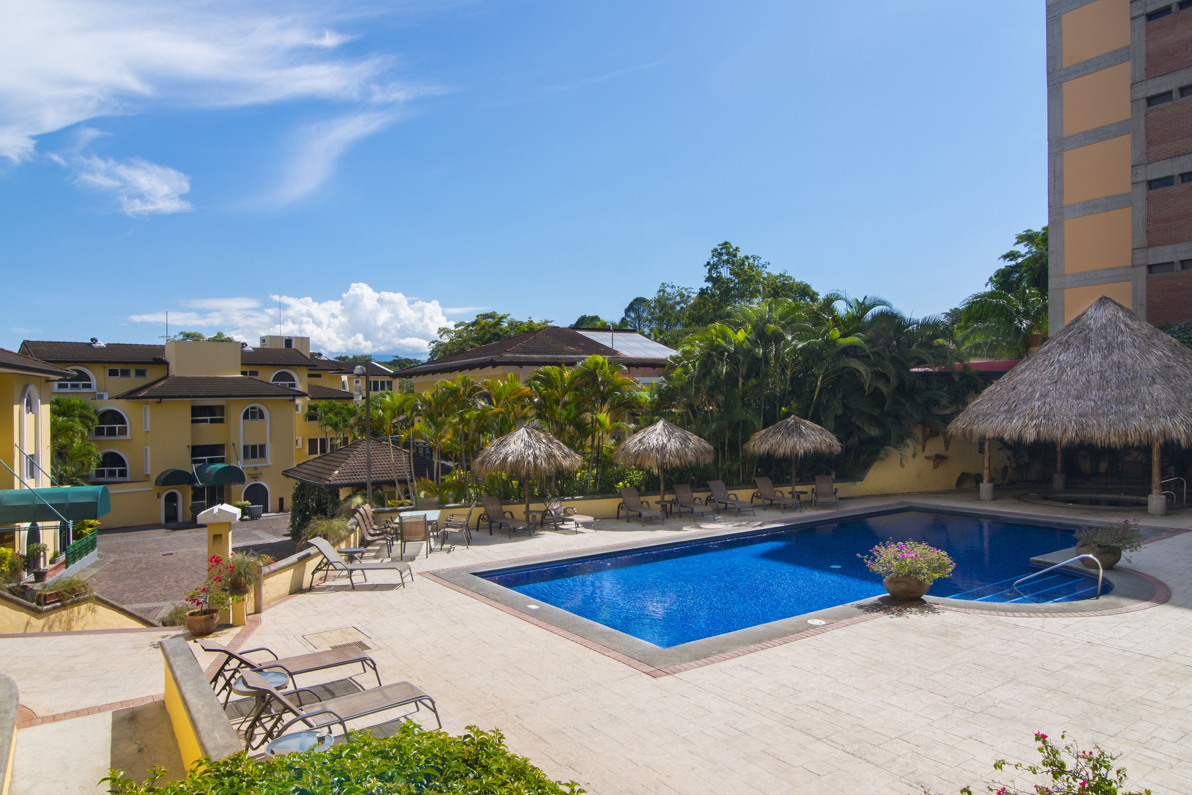 Piscina com espreguiçadeiras e guarda-sóis, rodeada por edifícios e vegetação.