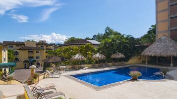 Piscina com espreguiçadeiras e guarda-sóis, rodeada por edifícios e vegetação.
