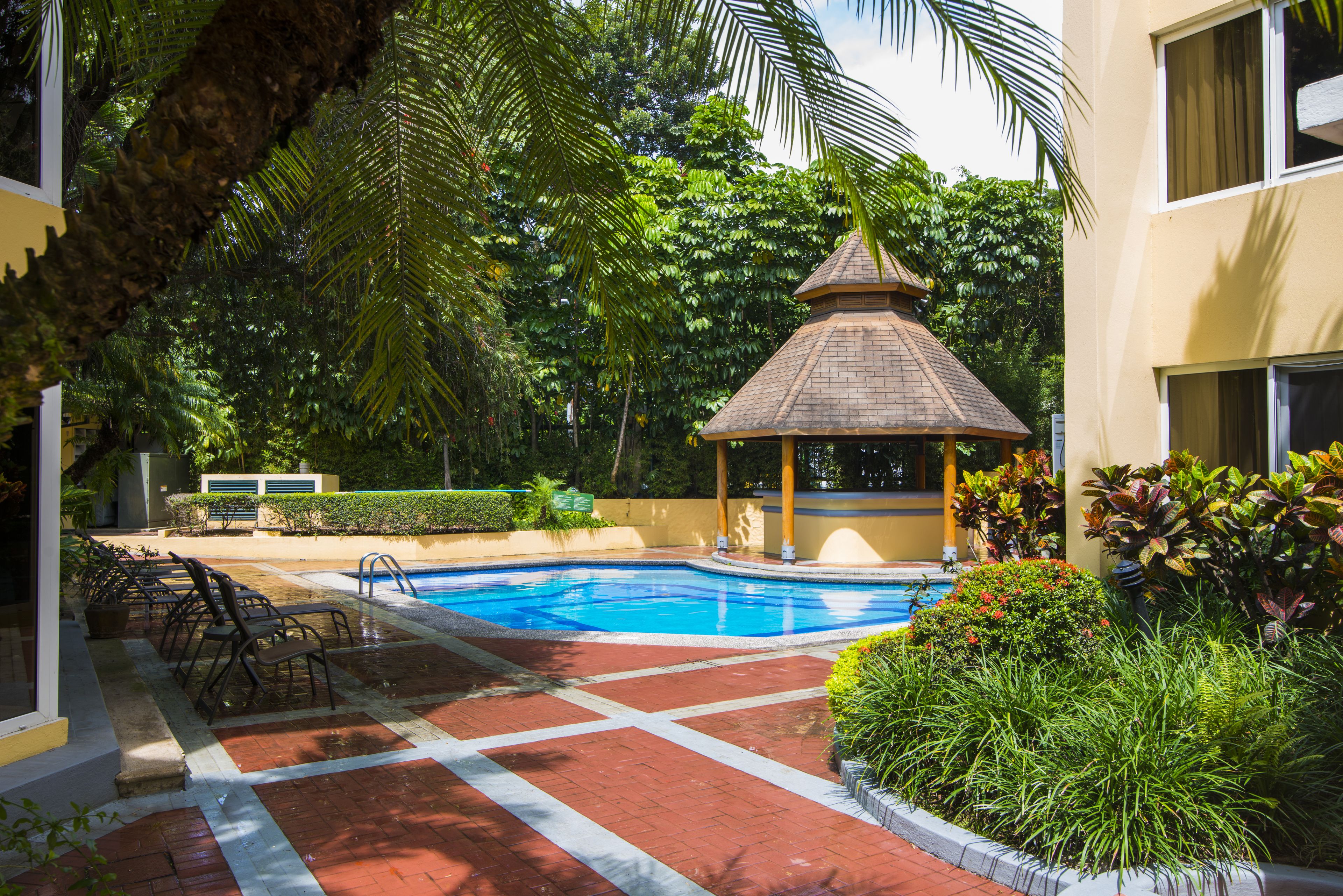 Una piscina rodeada de exuberante vegetación, un pabellón y un edificio con fachada amarilla.