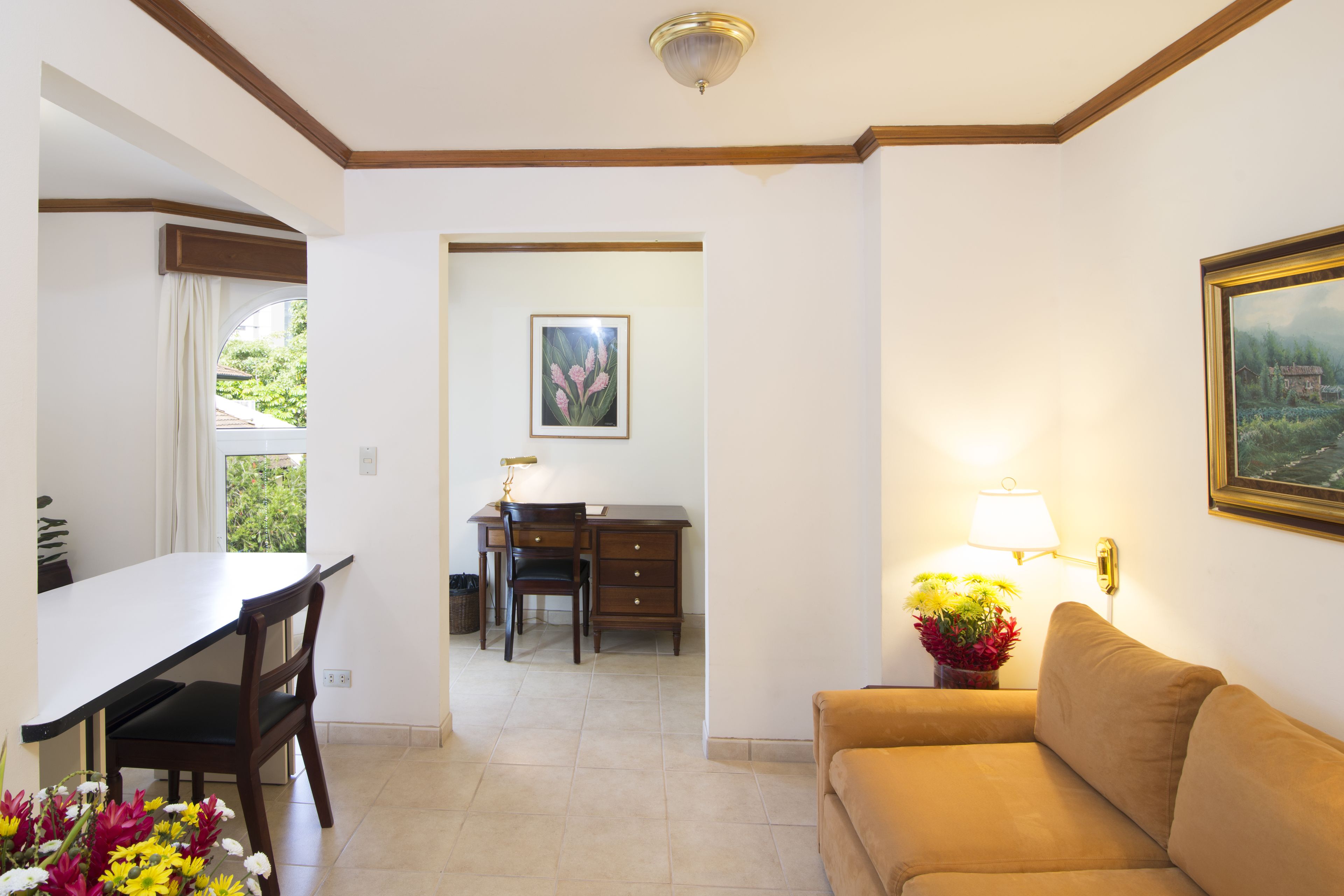A room with a dining table, chairs, a sofa, a desk, and a lamp.