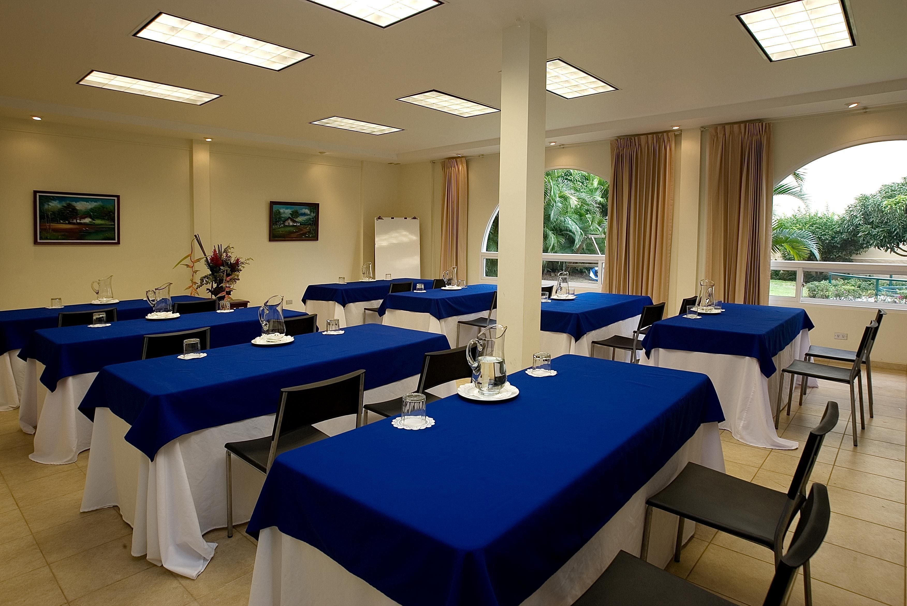 Un salón de conferencias con varias mesas cubiertas con manteles azules, fundas blancas para las sillas y equipado con vasos y jarras.