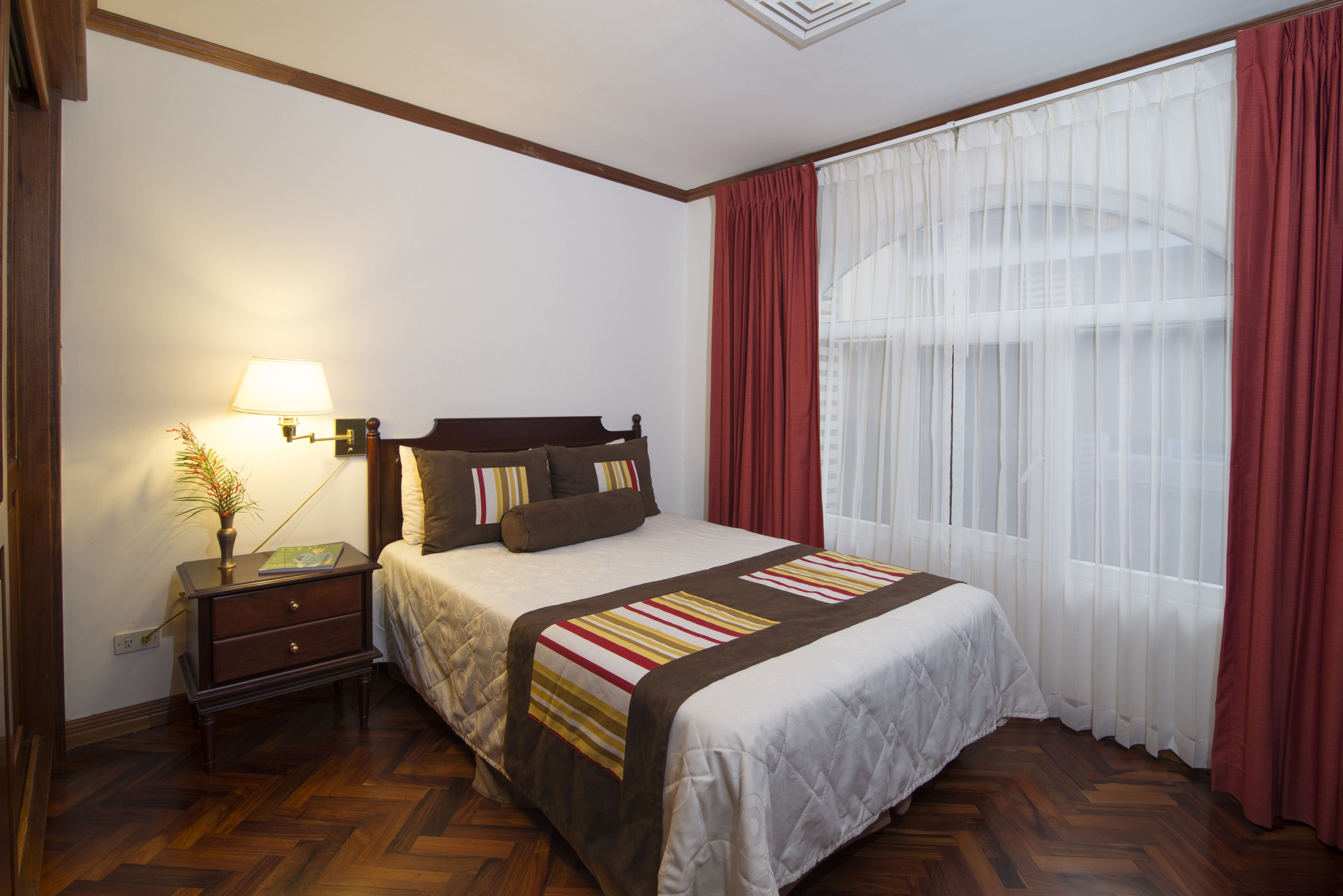 A neatly arranged bedroom with a bed, a bedside table with a lamp, and a window with sheer curtains.