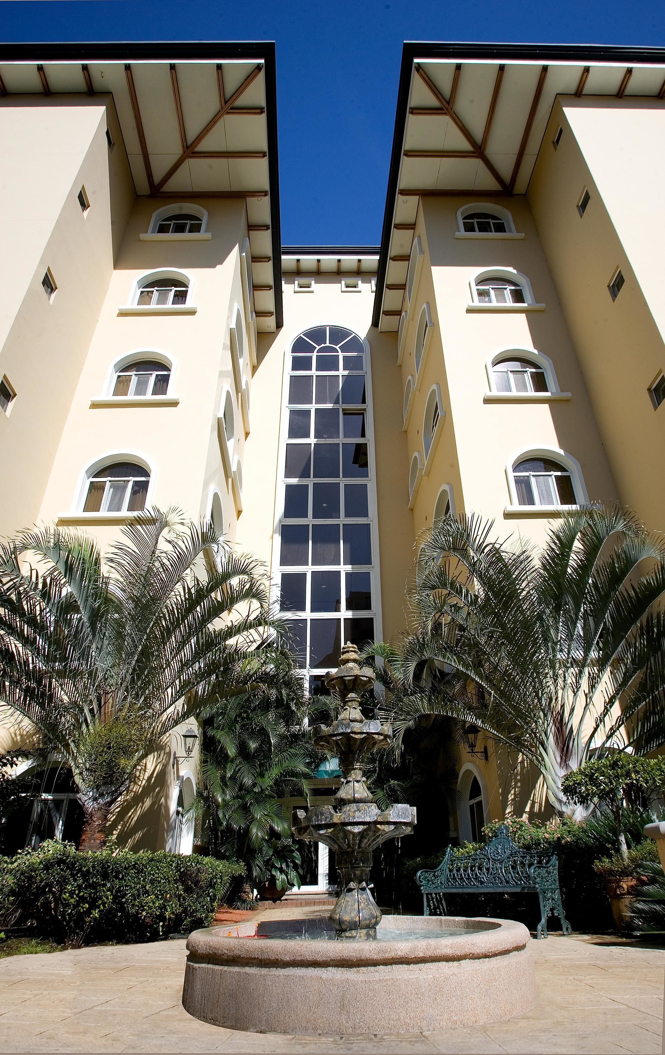 A building with a central arched window and multiple smaller windows, surrounded by palm trees and a fountain.