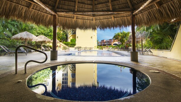 An infinity pool with a thatched roof, surrounded by lounge chairs and palm trees. - Apartotel & Suites Villas del Rio (San Rafael)
