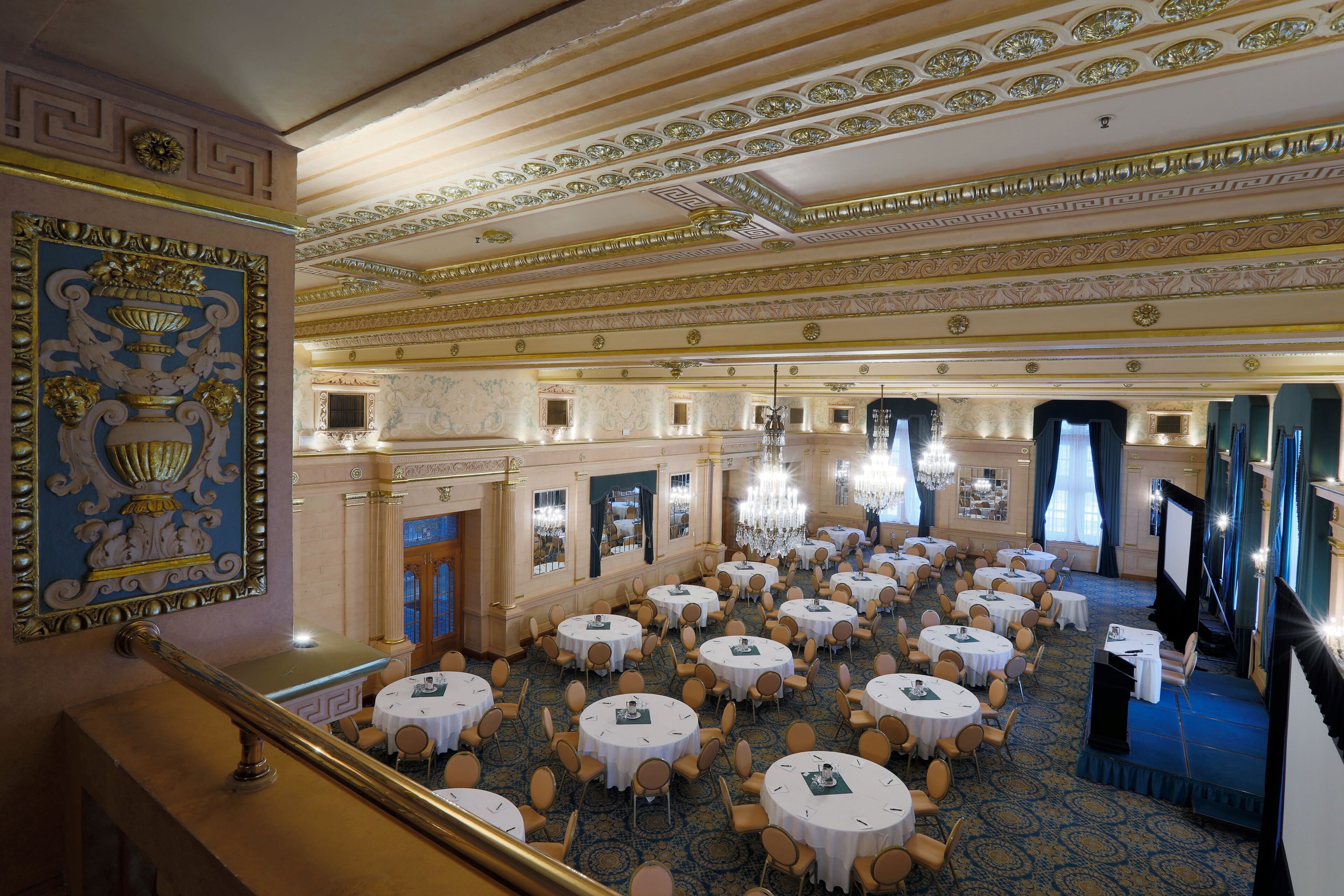 A grand ballroom with ornate ceiling decorations, multiple round tables, and a stage area.