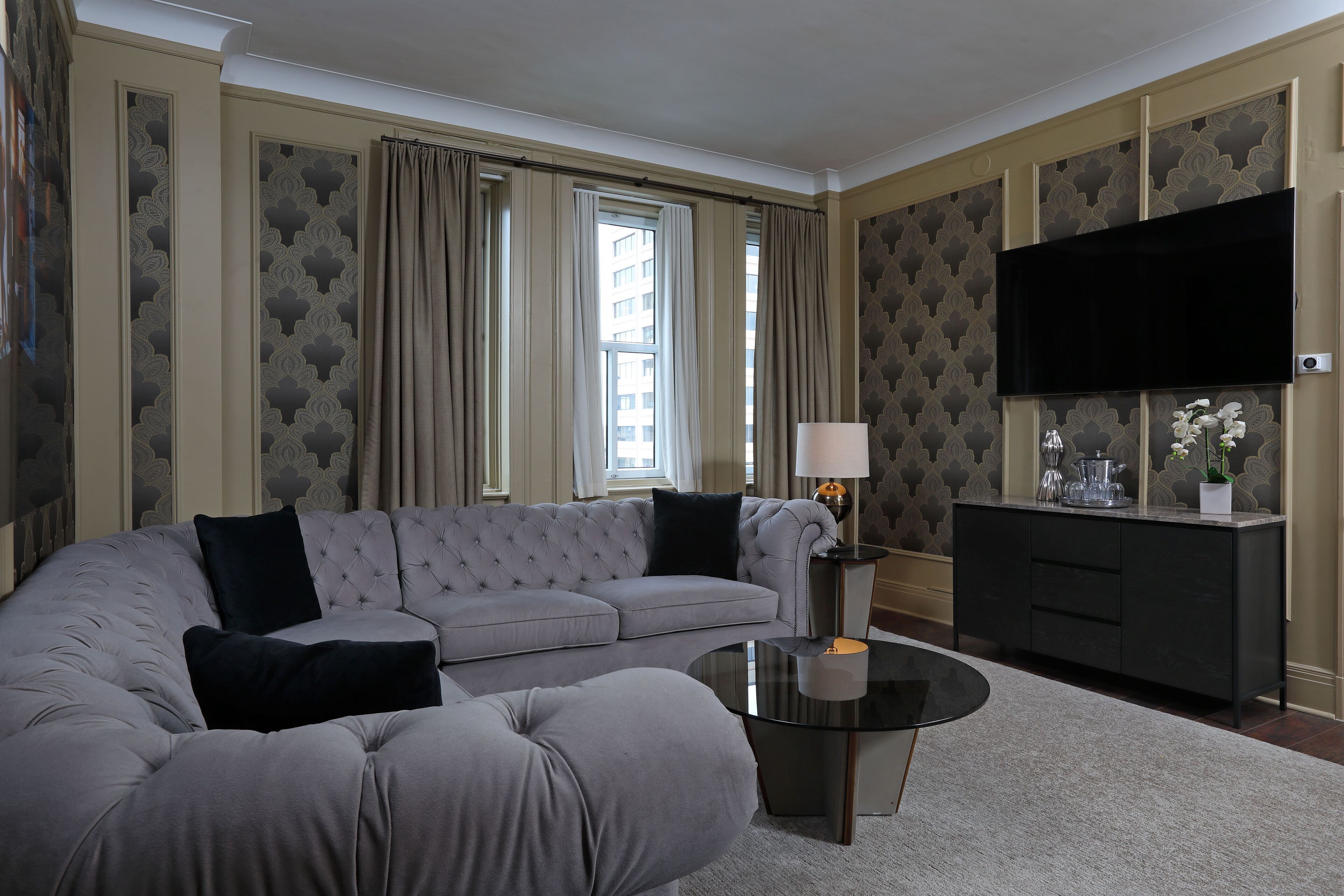 Un salon moderne avec un canapé d’angle gris, une table basse ronde, un téléviseur noir fixé au mur et un meuble décoratif.