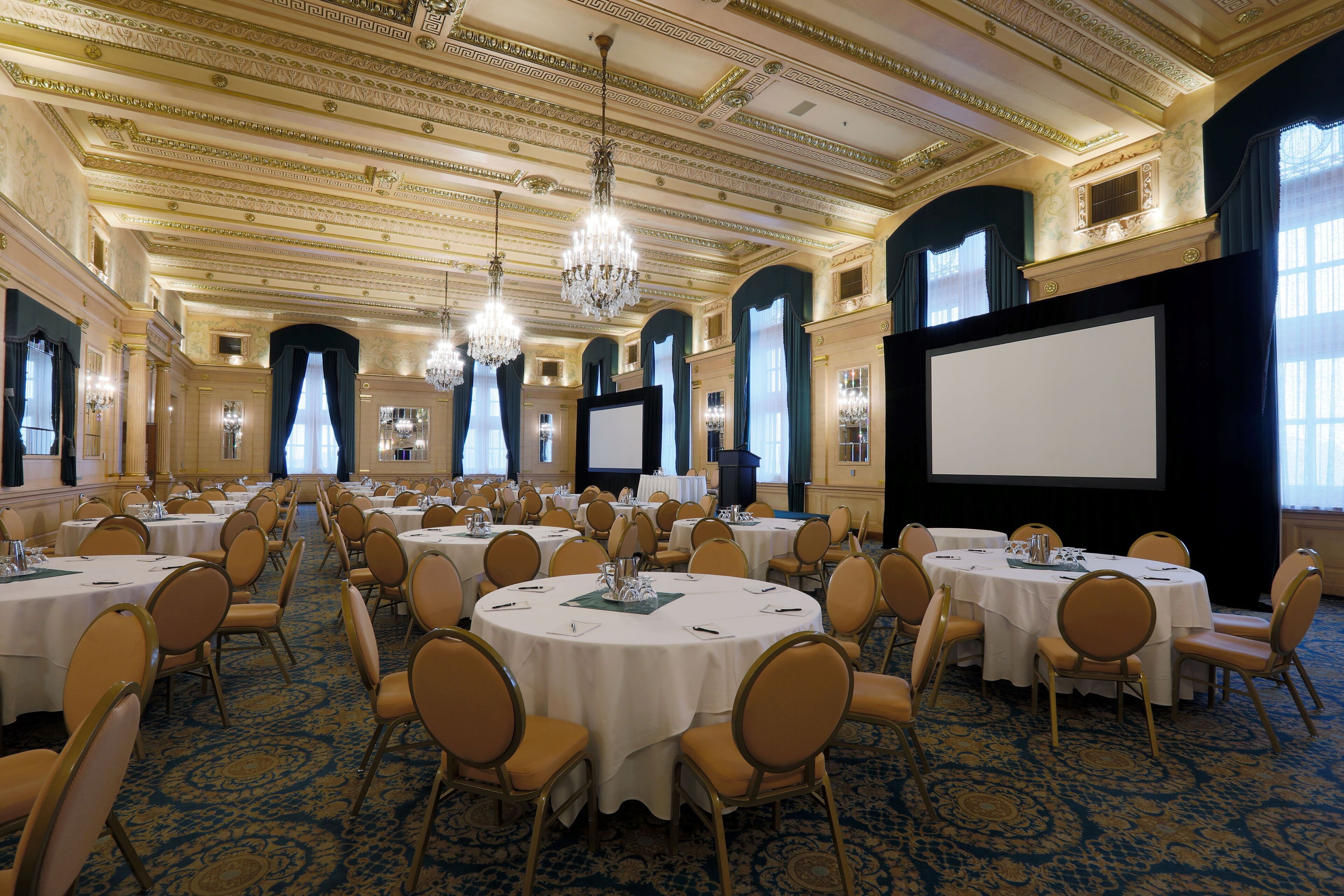 Un salón de banquetes con mesas redondas, sillas, candelabros y una gran pantalla.