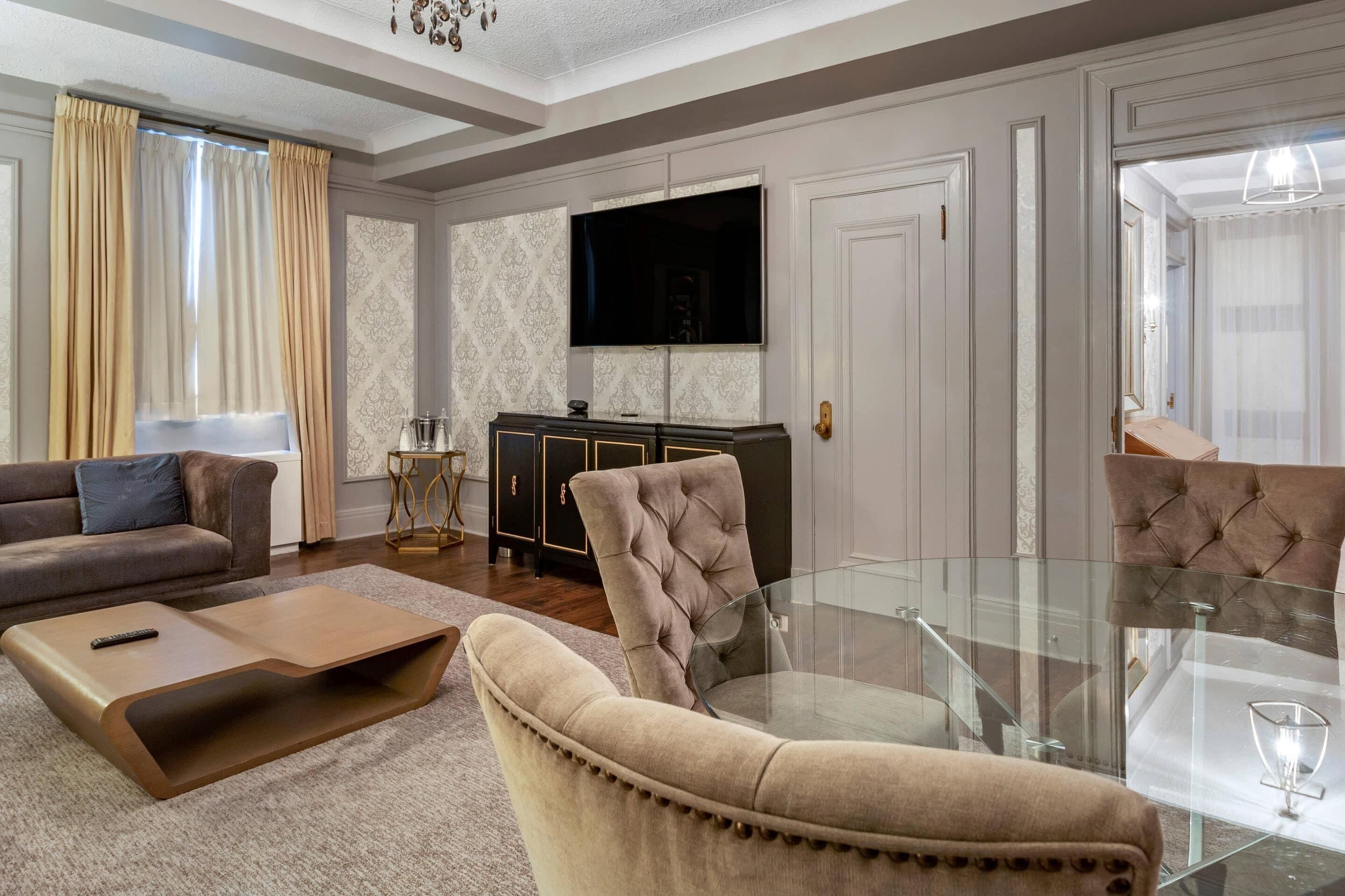 A living room with a glass-top coffee table, two upholstered chairs, a sofa, a television, and a cabinet.