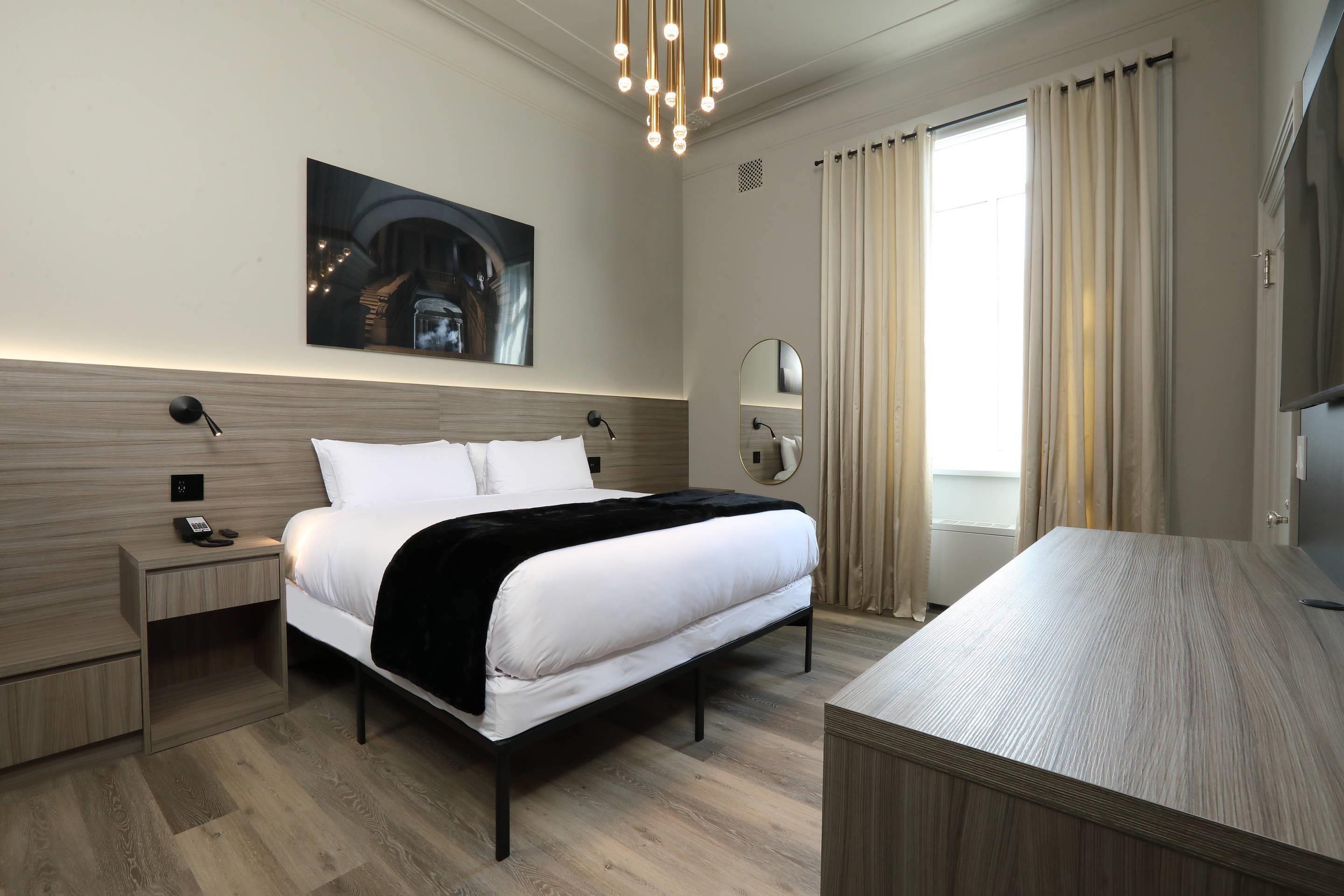 Quarto de hotel moderno com uma cama grande, mesas de cabeceira, escrivaninha, televisão de tela plana e uma janela ampla com cortinas.