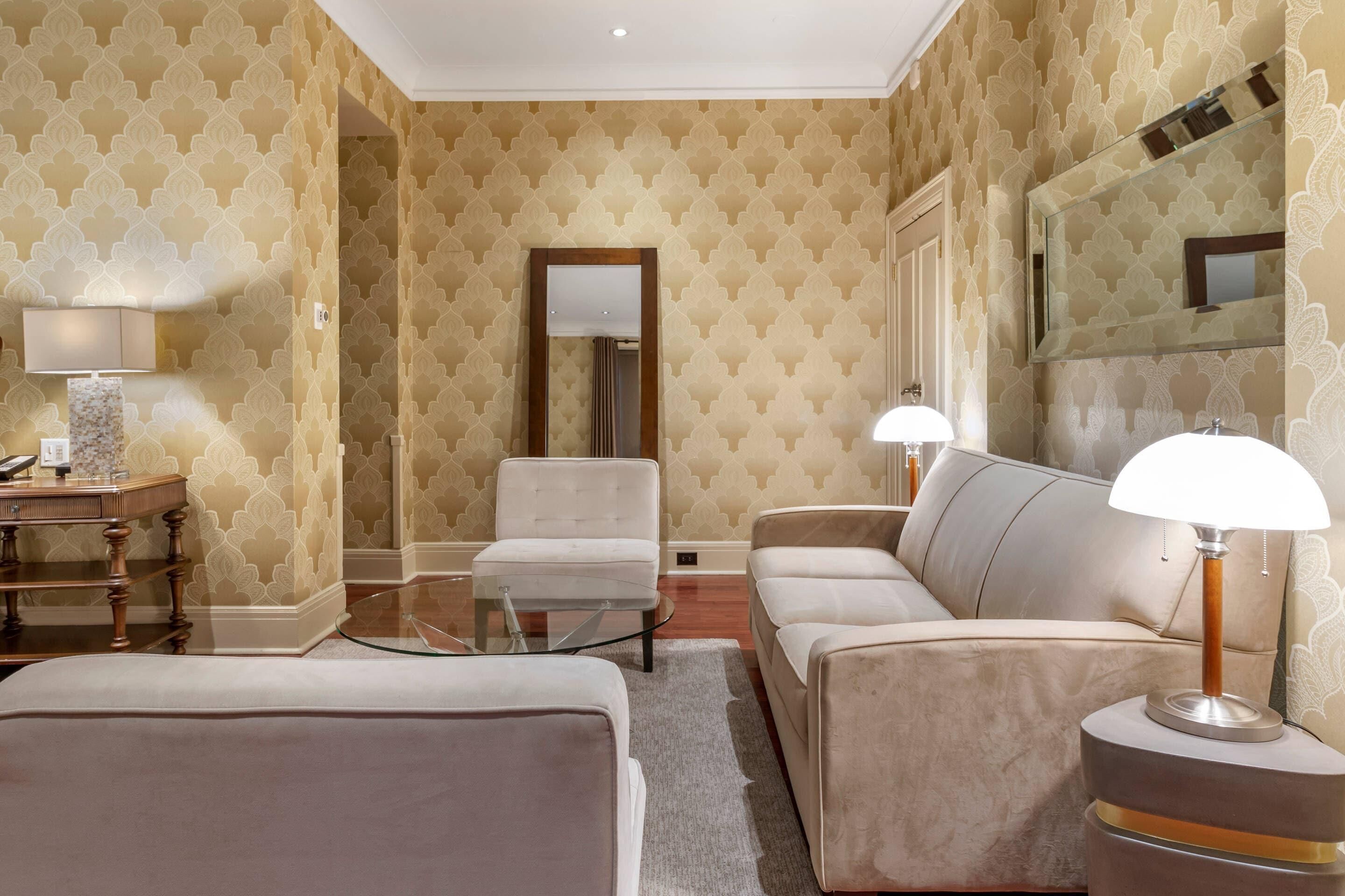 Habitación de hotel con sofá, sillón y una mesa de centro de vidrio. Se ve un espejo en la pared y una lámpara sobre una mesita.