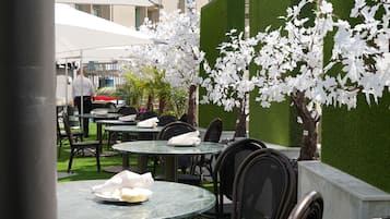 An outdoor seating area with round tables and chairs, surrounded by artificial grass and white flowering trees.