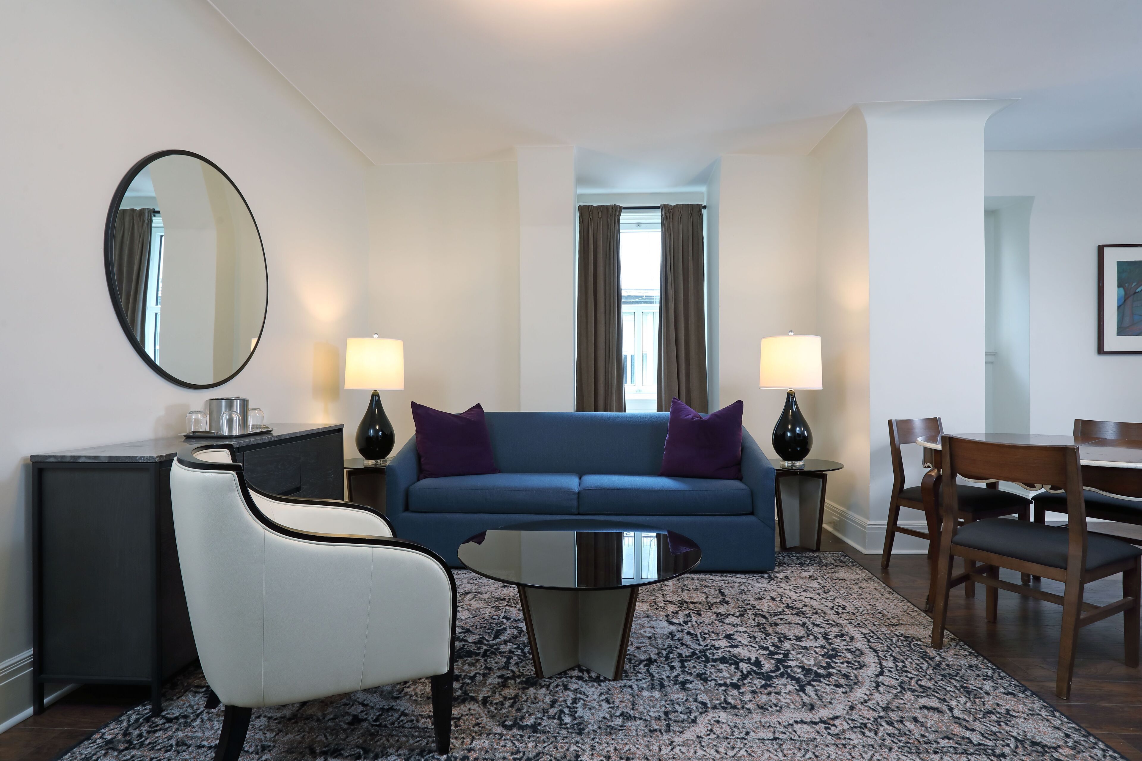 Un salon moderne avec un canapé bleu, un fauteuil blanc et une table basse ronde. On y trouve deux lampes, une table à manger avec des chaises et un grand miroir accroché au mur.