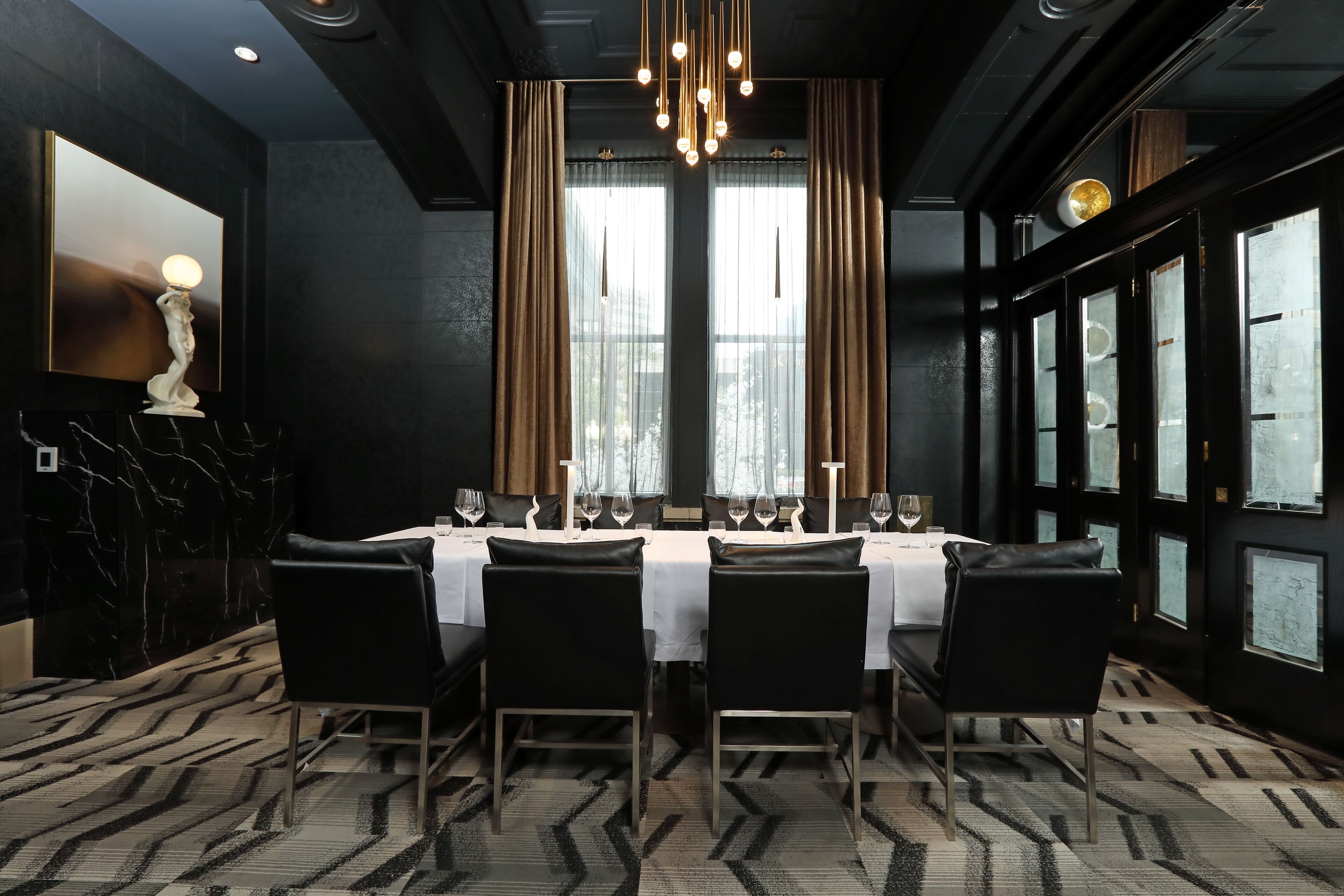 A dining area with a table set for four, black chairs, a chandelier, and large windows with curtains.
