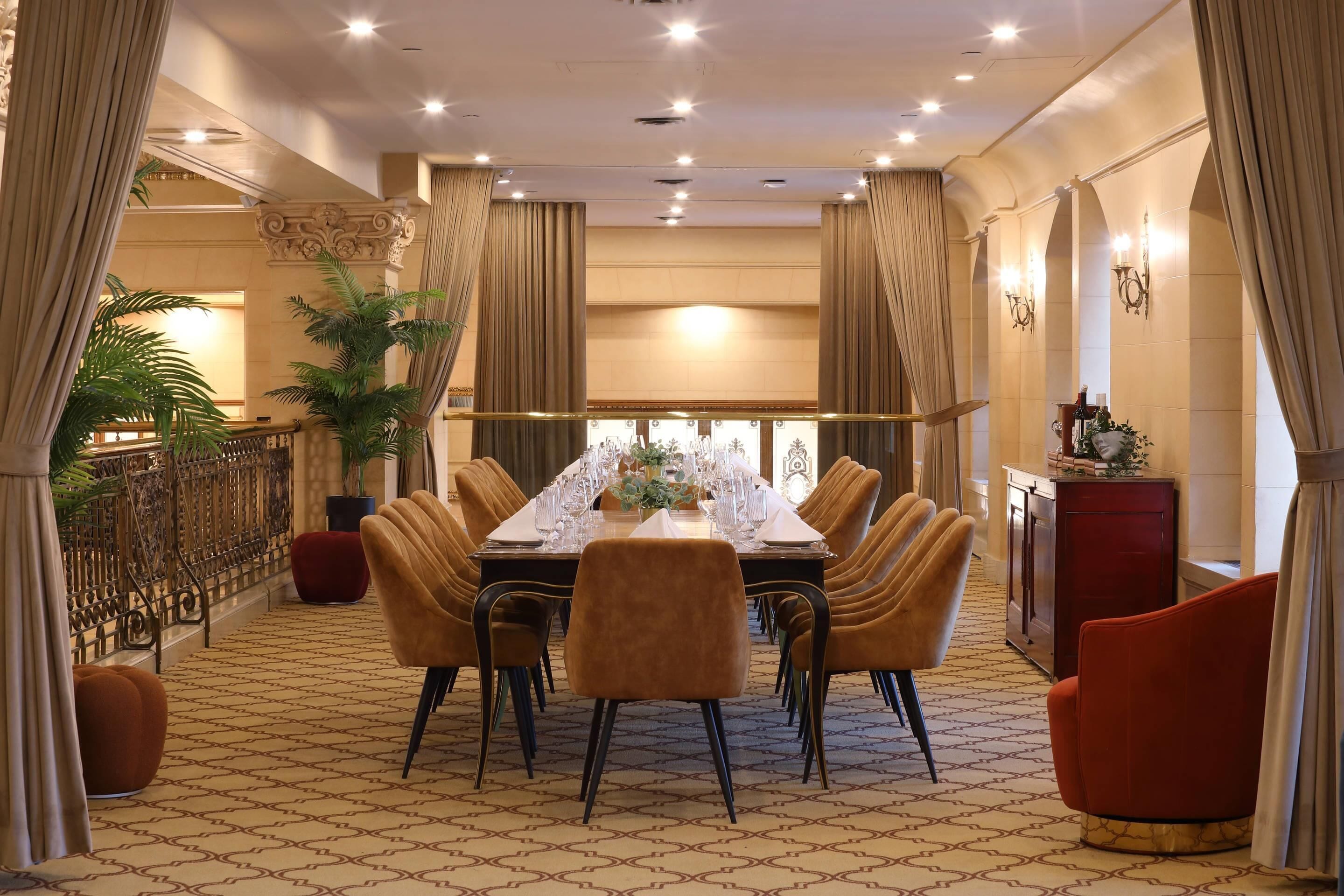 A dining area with a long table set for a meal, surrounded by chairs, with a potted plant and a red cabinet in the background.