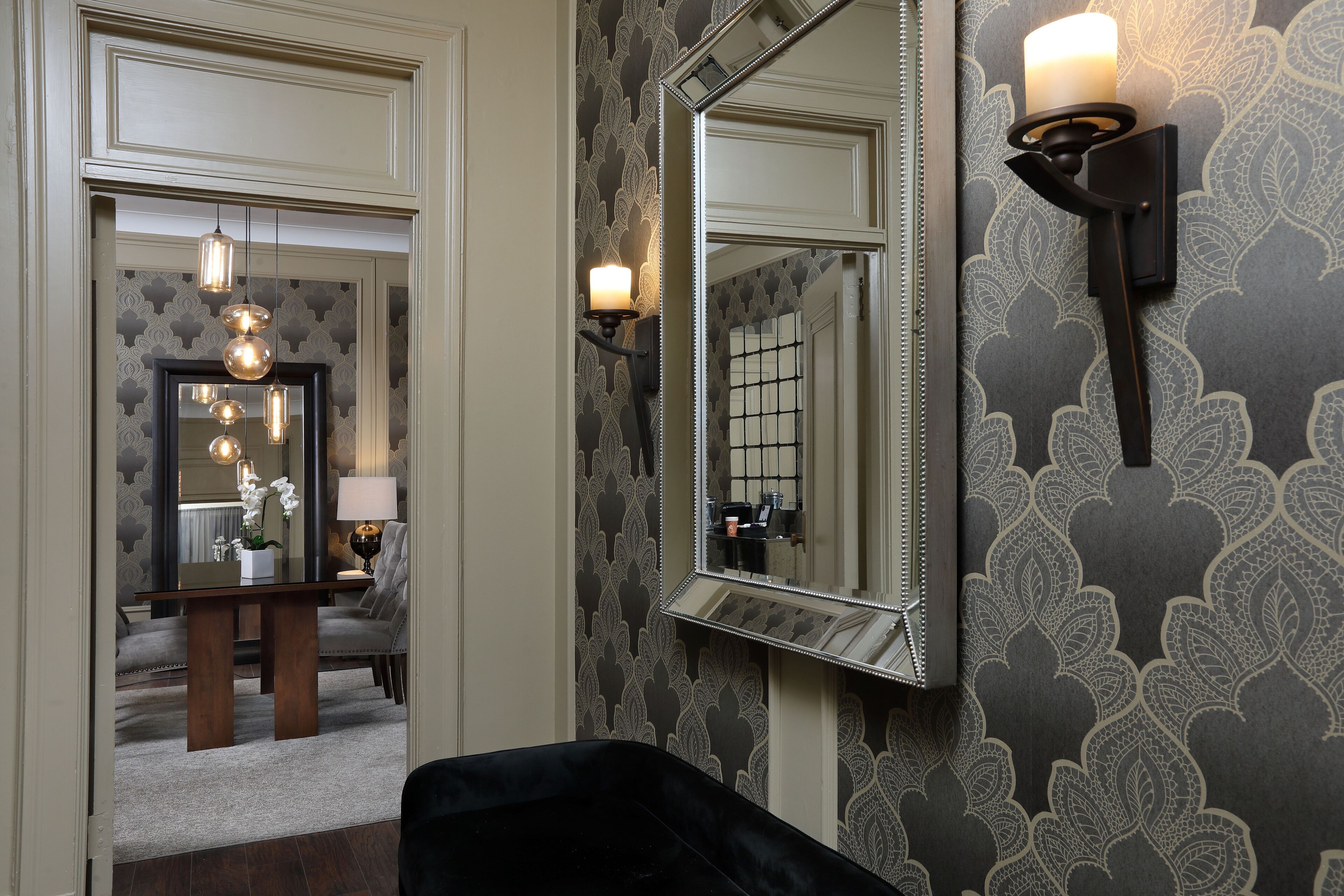 A room with a mirror, wall sconces, a table with a vase, and a chair.