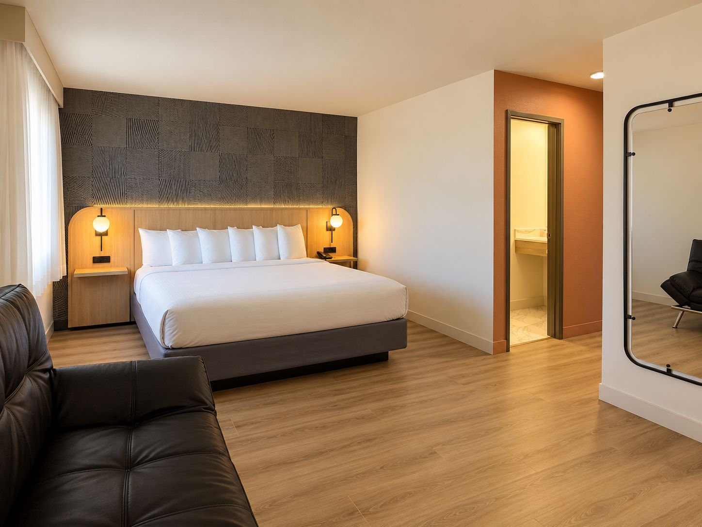 A modern hotel room with a large bed, bedside lamps, a wooden headboard, a leather sofa, and a mirror.