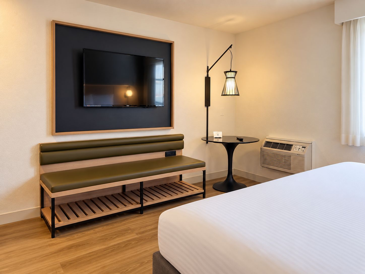 A hotel room with a bed, a bench, a TV mounted on the wall, a table with a lamp, and an air conditioning unit.