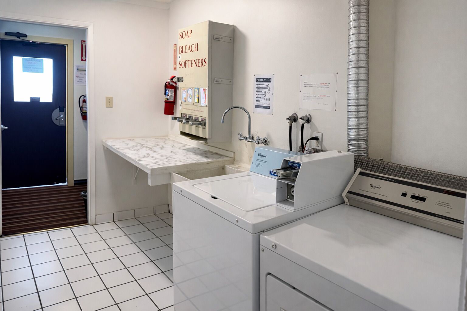 A laundry room with a sink, a washing machine, and a dryer. There is a fire extinguisher and a sign for soap, bleach, and softeners.