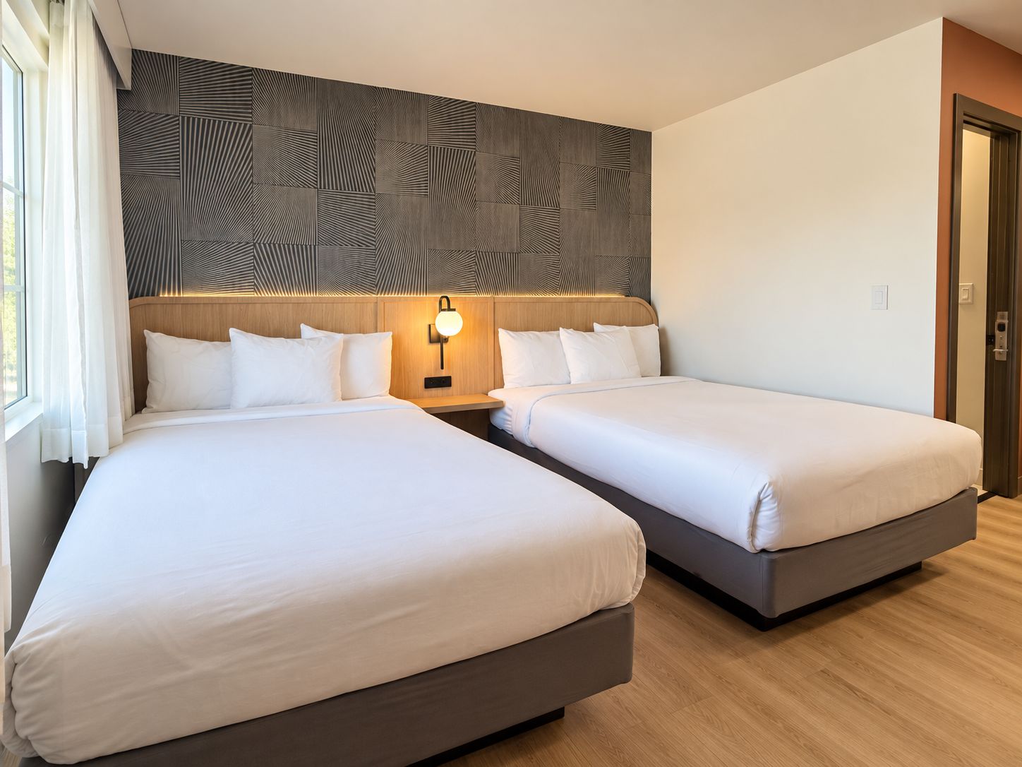 Habitación de hotel con dos camas, cabecera de madera, lámpara de pared y ventana con cortinas.