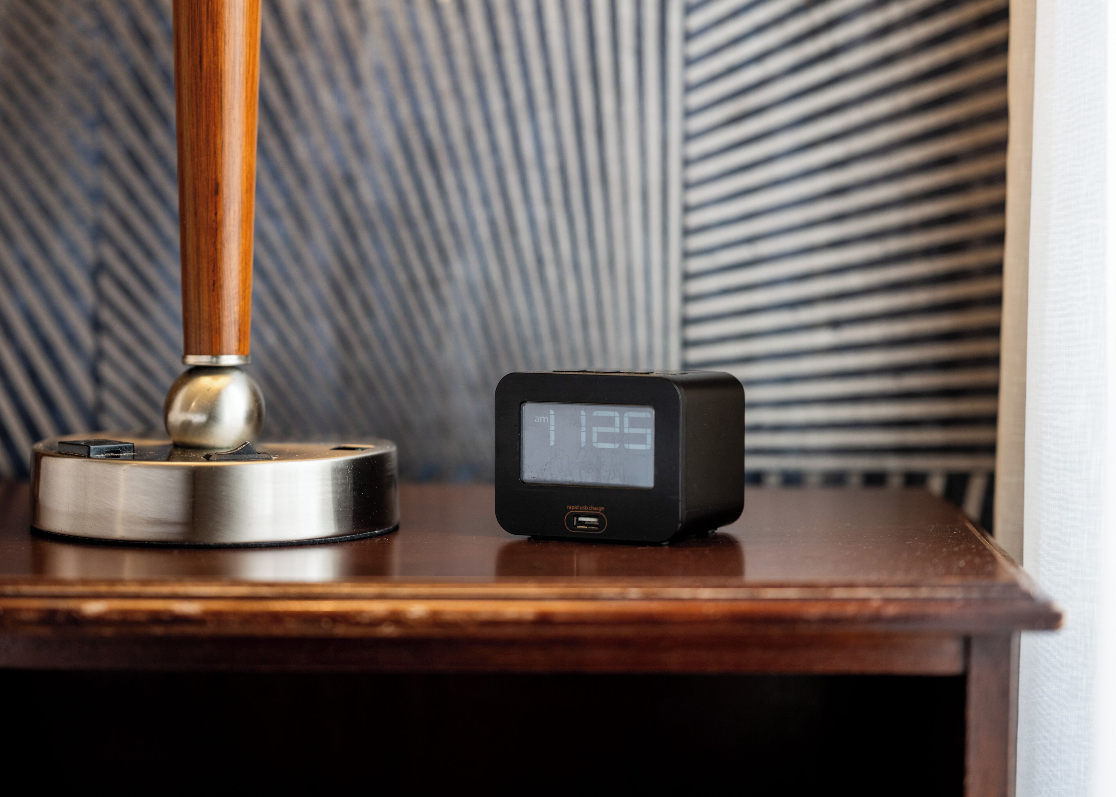 Um relógio digital mostra 12:25 sobre uma mesa de madeira, ao lado de um abajur com base metálica.