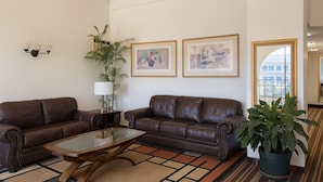 A living room with two brown leather sofas, a glass coffee table, a potted plant, and two framed artworks on the wall.