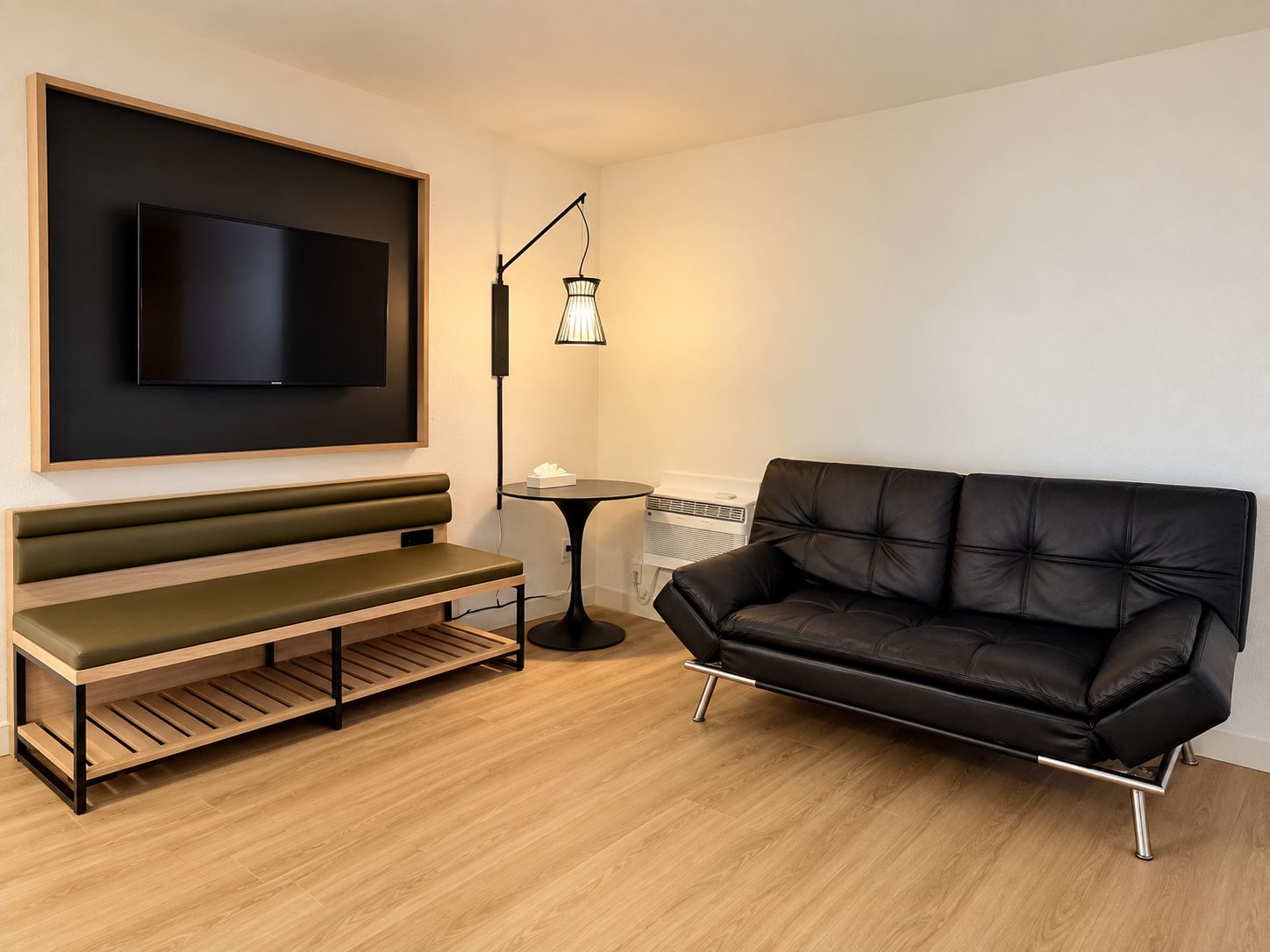 A modern living room with a black leather sofa, a wooden bench, a flat-screen TV mounted on a wooden frame, a floor lamp, and a small table with a tissue box.