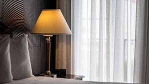 A hotel room with a bed, a bedside table with a lamp, a clock, and sheer curtains.