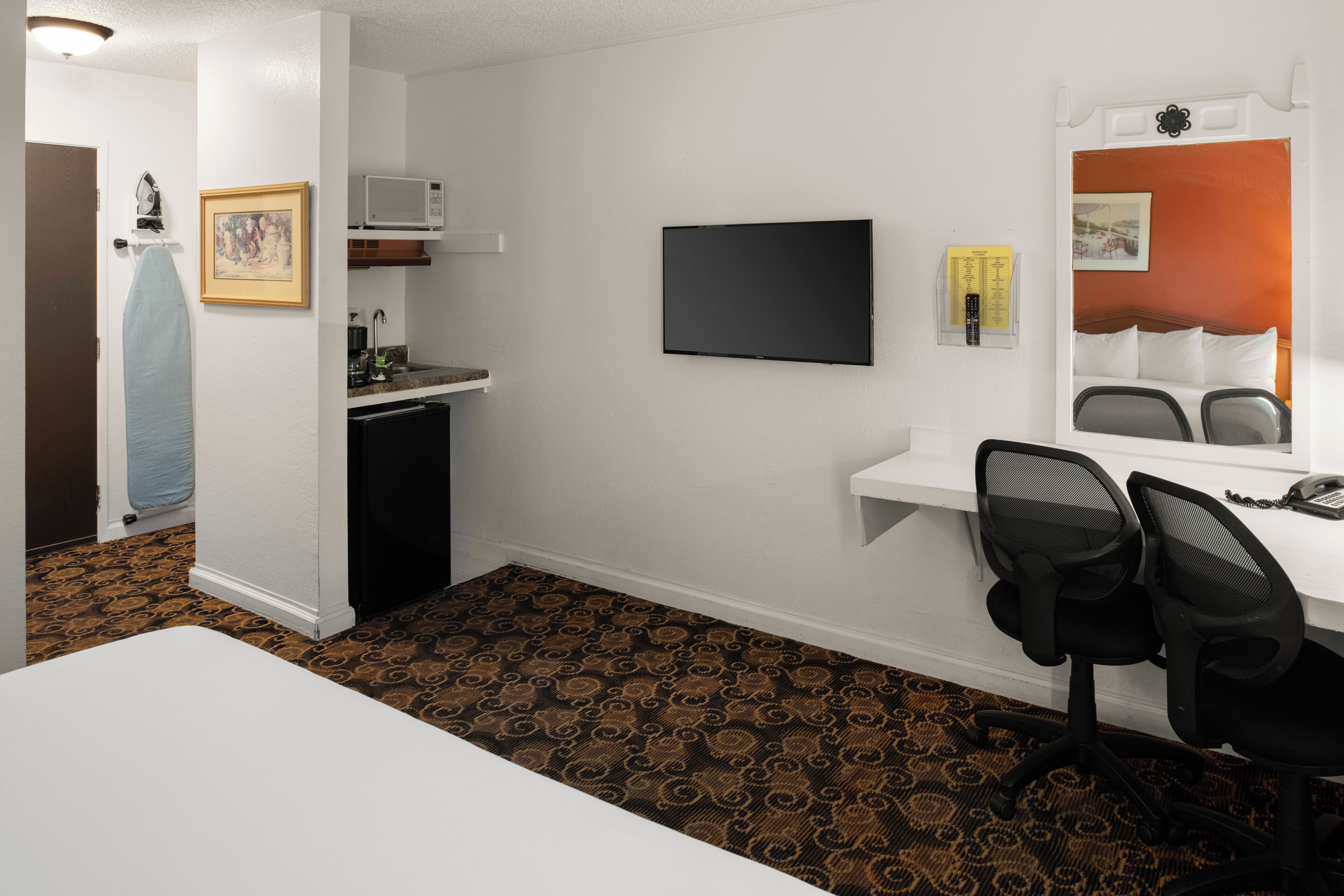 A hotel room with a kitchenette, a flat-screen TV, a desk with two chairs, a patterned carpet, and a bed with a headboard.