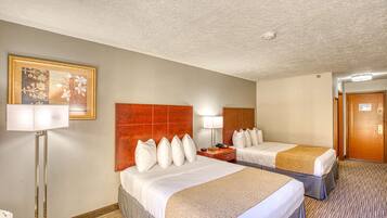 Habitación de hotel con dos camas, un cuadro grande enmarcado en la pared, una lámpara de pie y una puerta.
