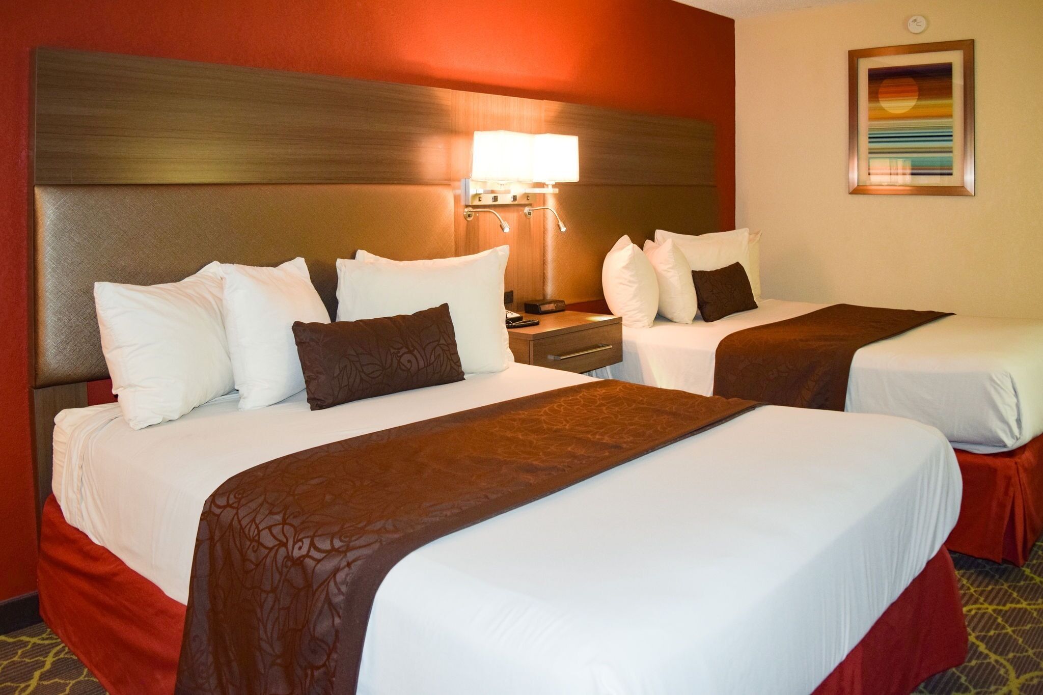Una habitación de hotel con dos camas, cada una con ropa blanca y mantas marrones. Entre las camas hay una mesita de noche con una lámpara. La habitación tiene una alfombra roja y un cabecero de madera.