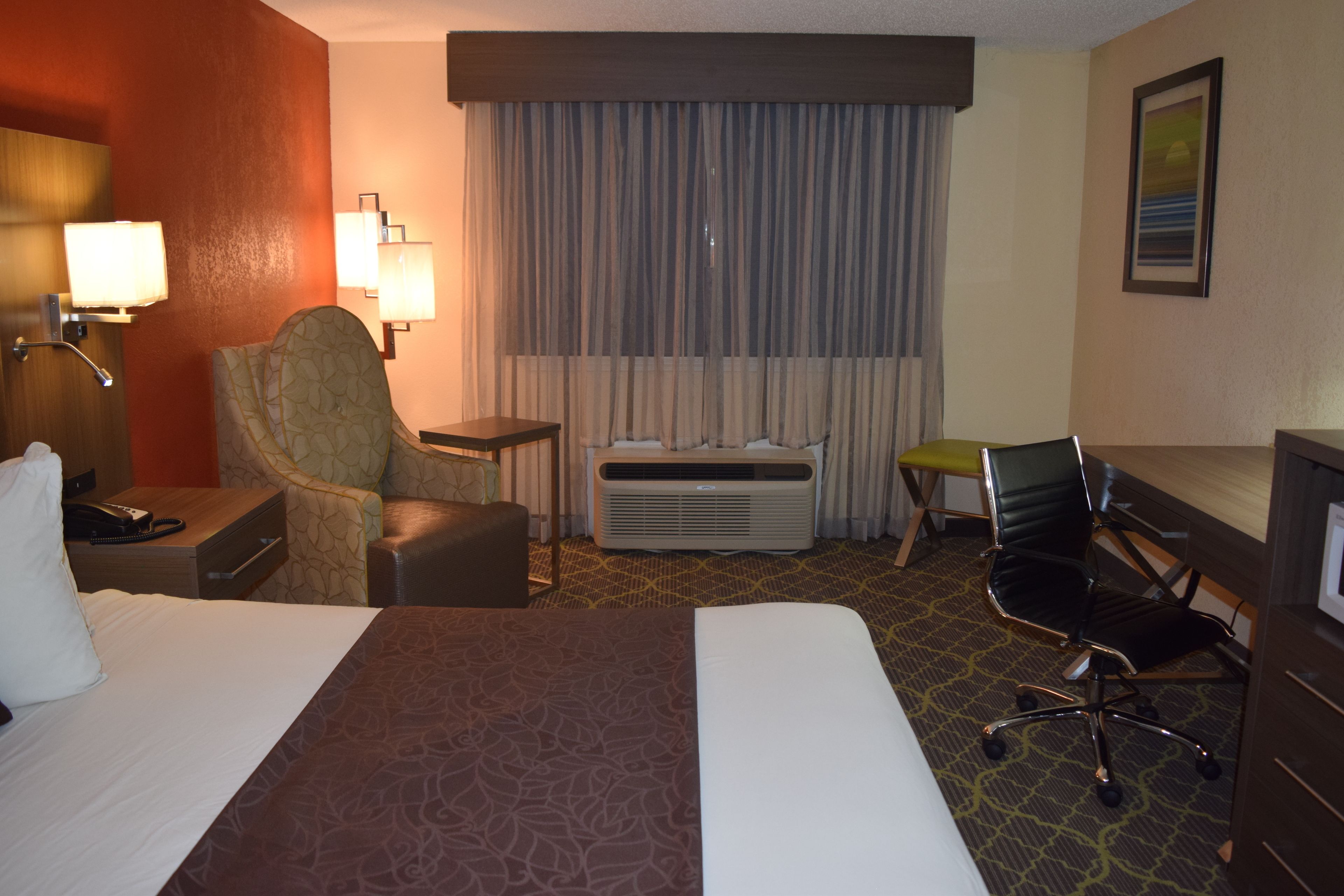 A hotel room with a large bed, two armchairs, a desk, a chair, an air conditioning unit, and a window with curtains.