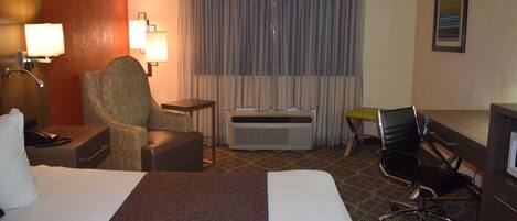 A hotel room with a large bed, two armchairs, a desk, a chair, an air conditioning unit, and a window with curtains.