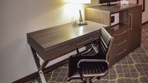 A hotel room with a wooden desk, a black office chair, a lamp, a flat-screen TV, and a minibar.