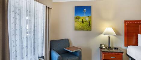 A hotel room with a bed, a chair, a nightstand with a lamp, a wall-mounted painting, and a window with curtains.