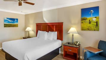 A hotel room with a large bed, two bedside tables with lamps, a small table, a blue chair, a ceiling fan, and two wall-mounted paintings.