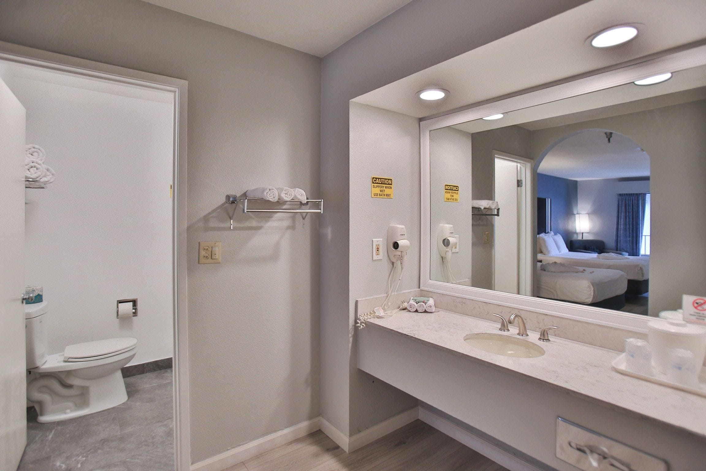 A bathroom with a toilet, sink, and mirror. There are towels on a shelf and a caution sign on the wall.