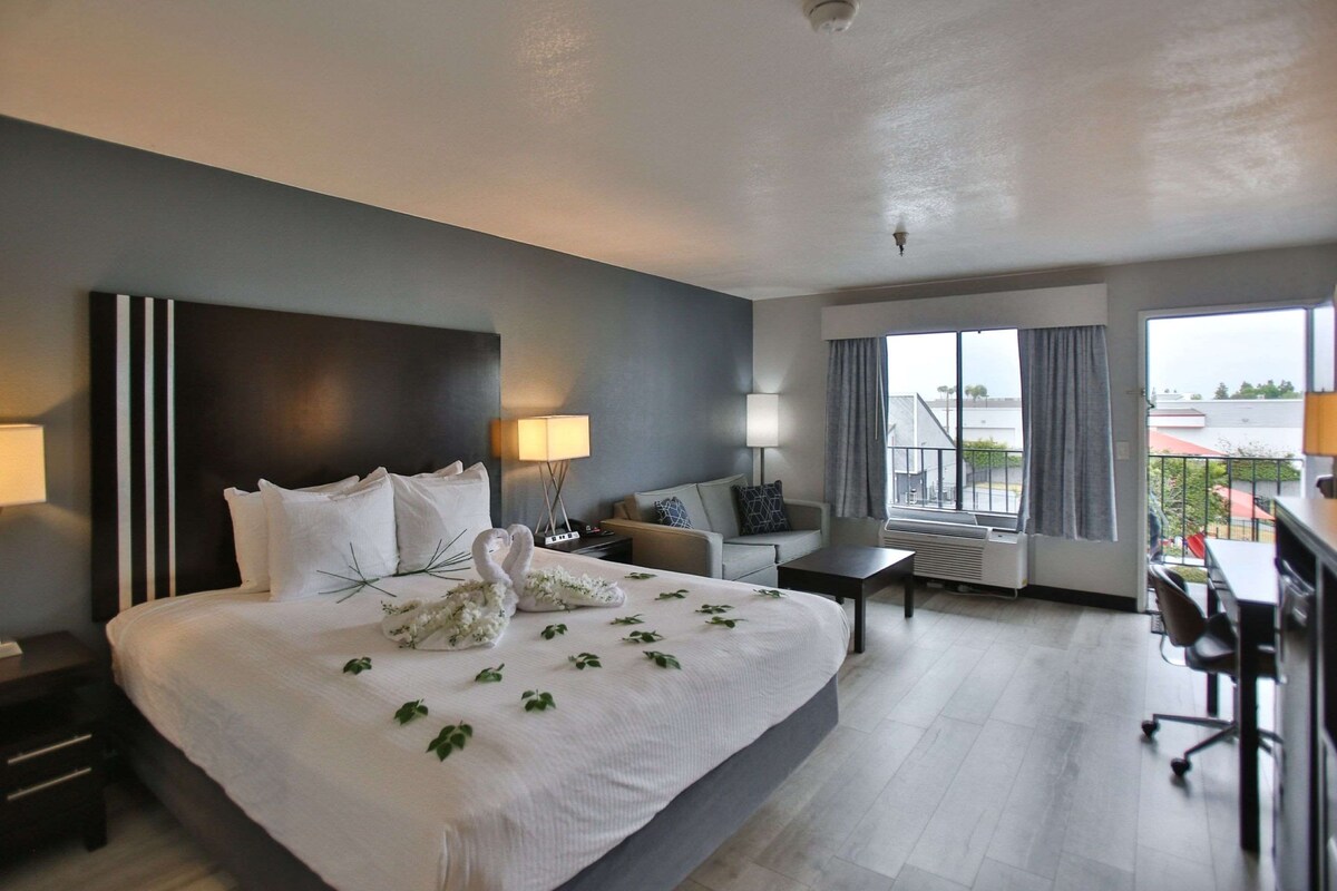 A hotel room with a large bed, a desk, a chair, a sofa, and a balcony with a view.