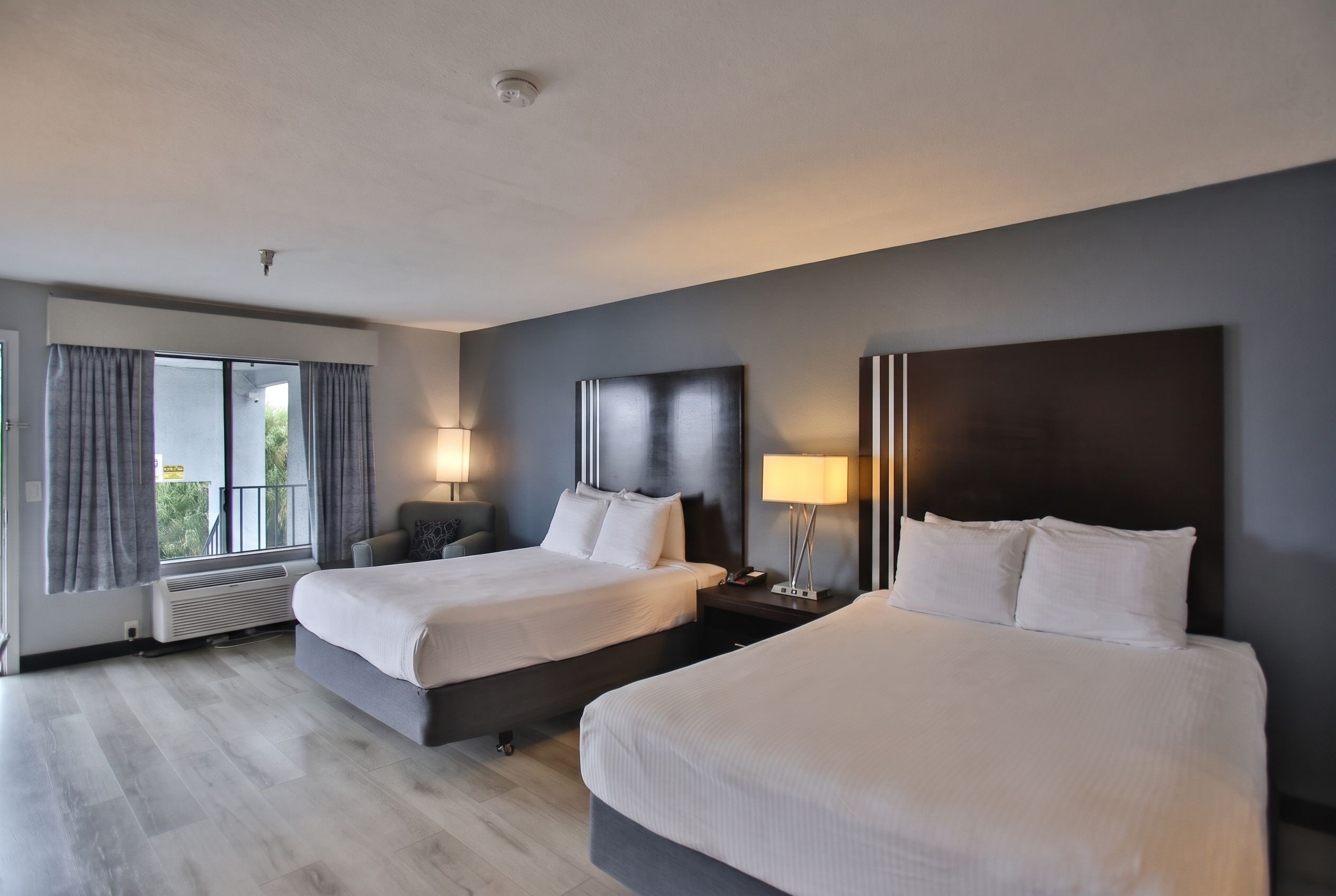 Une chambre d’hôtel avec deux lits, une grande fenêtre avec des rideaux, une table de chevet avec une lampe et un radiateur fixé au mur.