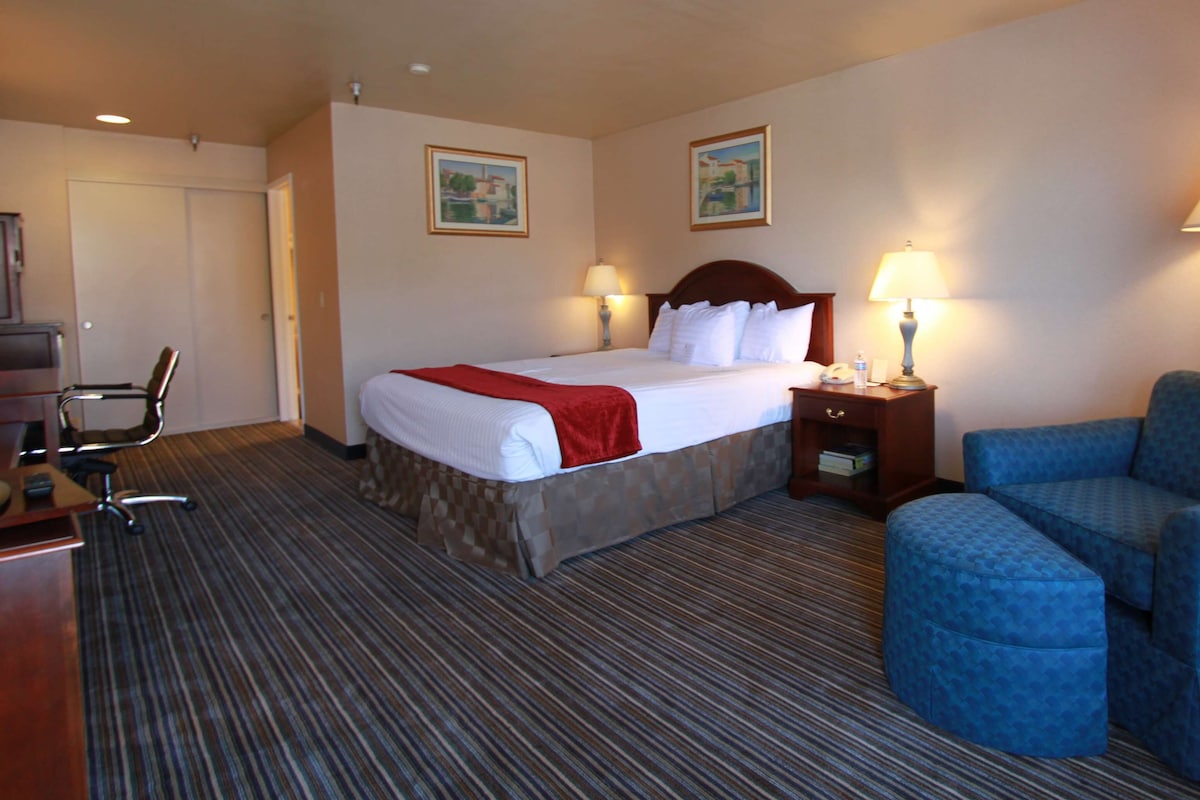 A hotel room with a bed, two bedside lamps, a desk with a chair, and a blue armchair.
