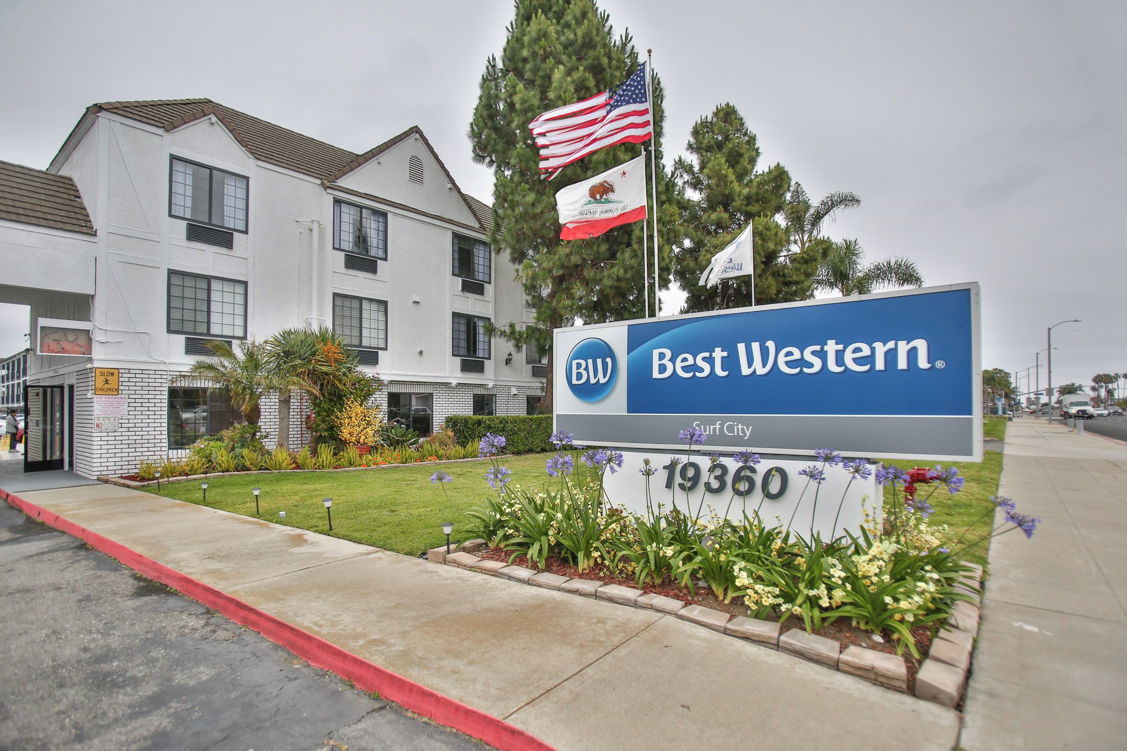 A Best Western hotel sign with the address 19360, surrounded by a landscaped garden with flowers and a flagpole with two flags.