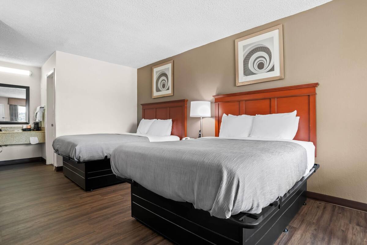 A hotel room with two beds, a wooden floor, a bathroom with a mirror, and two framed pictures on the wall.