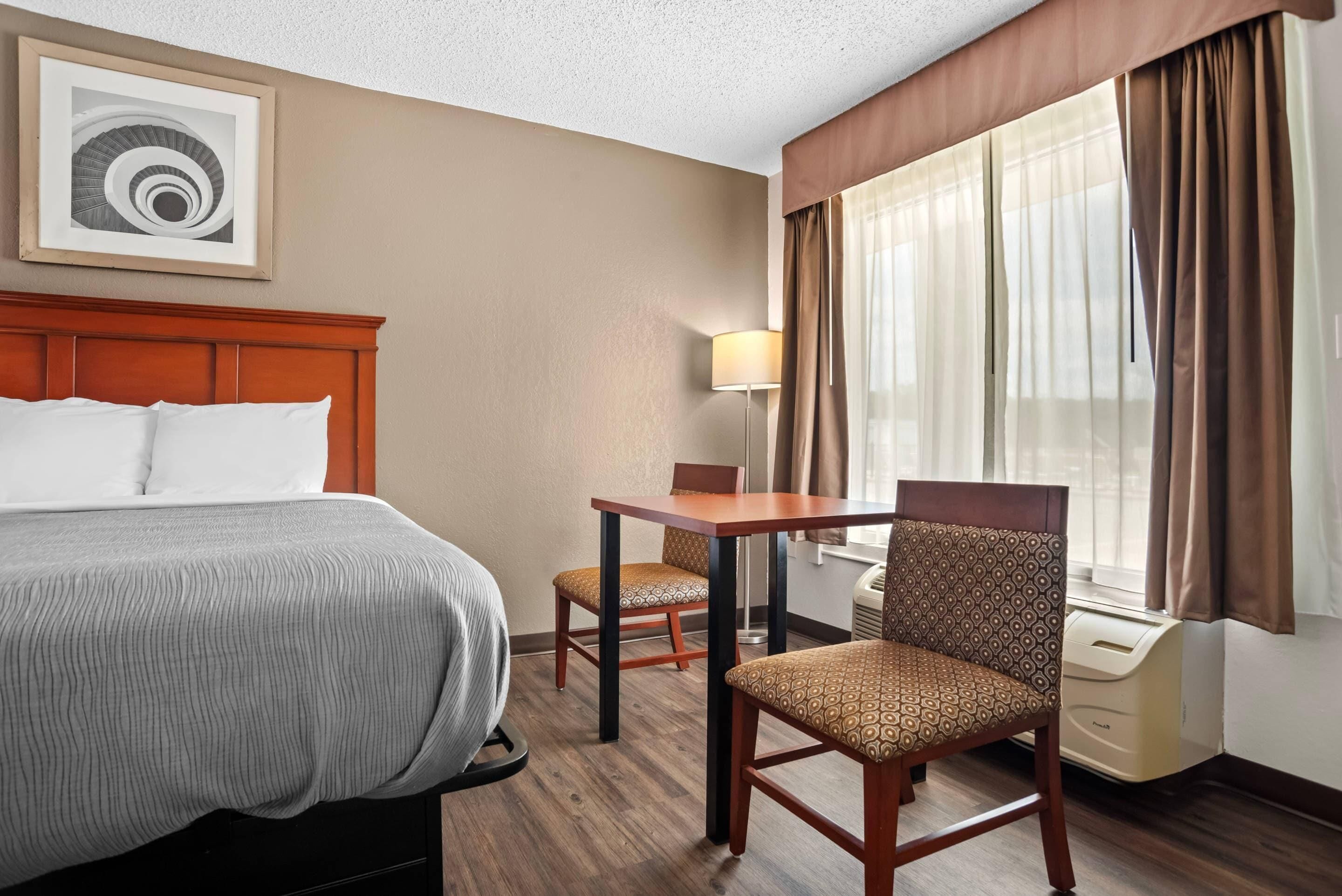 A hotel room with a bed, a desk, two chairs, a lamp, and a window with curtains.
