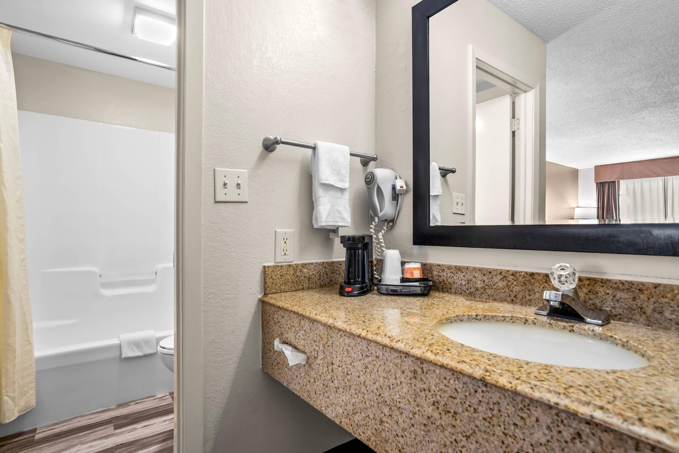 Une salle de bain dotée d’un comptoir en granit, d’un lavabo, d’un miroir, d’un porte-serviettes, d’un sèche-cheveux et d’une machine à café.