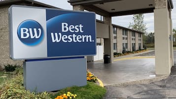 Señal de Best Western con un logotipo azul y blanco, un edificio con acceso cubierto y un estacionamiento con líneas amarillas.