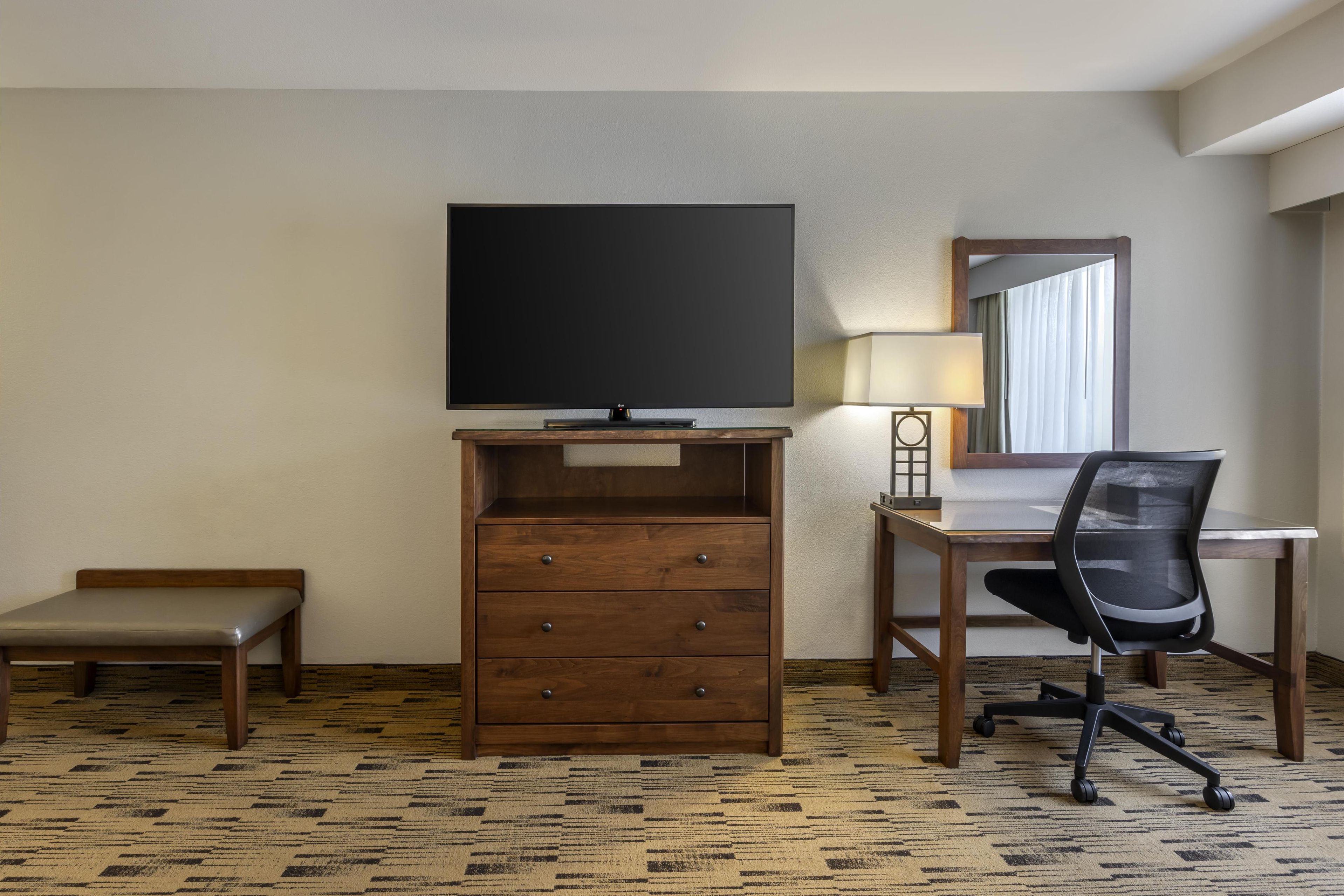 A hotel room with a wooden desk, a television, a chair, a small bench, and a lamp.