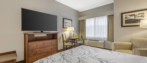 A hotel room with a large bed, a TV, a desk with two chairs, a window with curtains, and a wall-mounted air conditioner.
