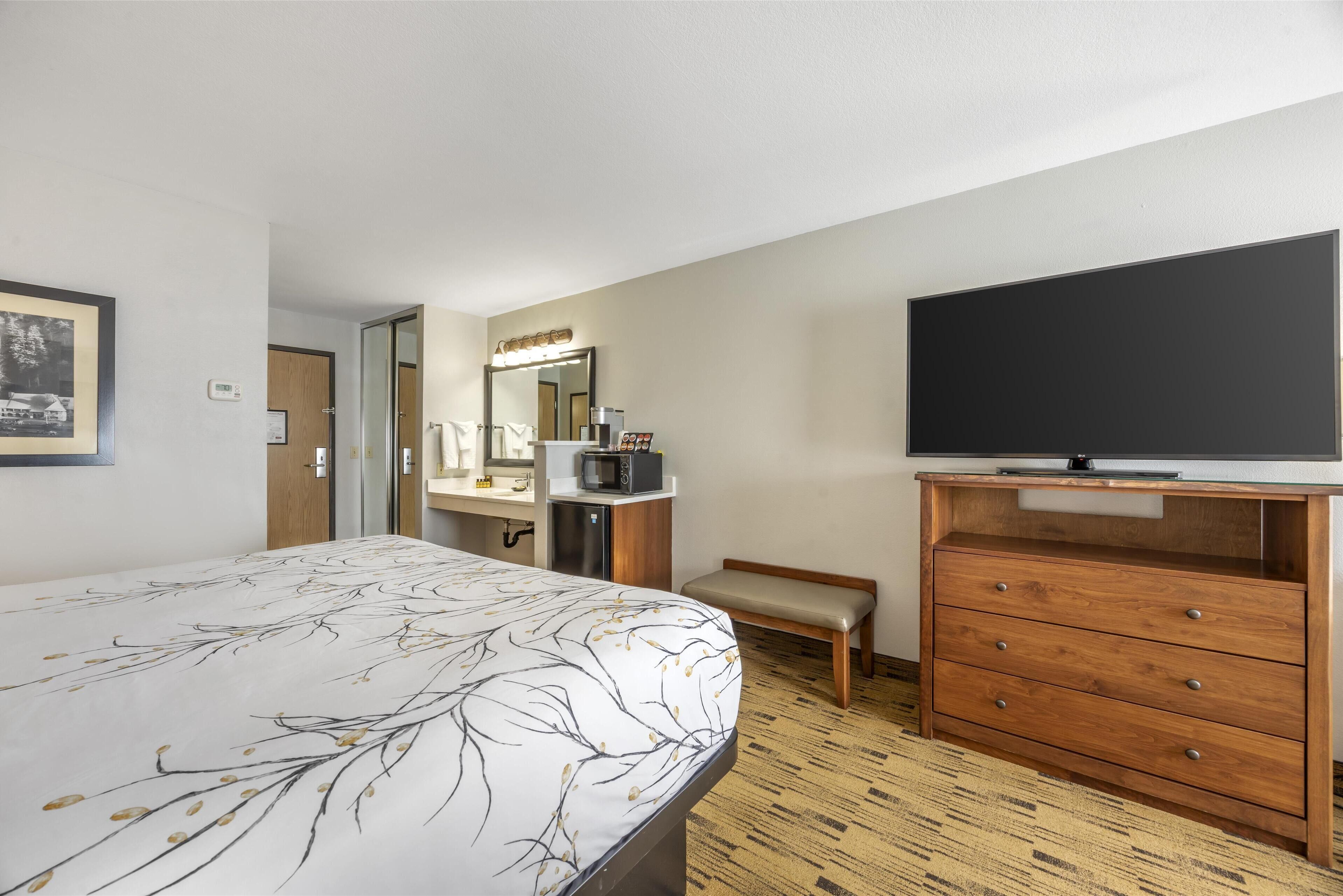 A hotel room with a bed, a television, a wooden dresser, a bench, a microwave, and a mirror.