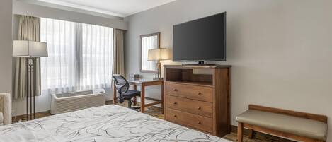 A hotel room with a large bed, a desk with a chair, a television, a bench, and a window with curtains.