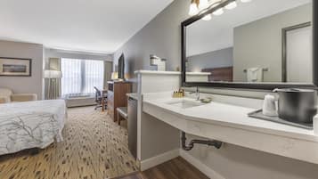 A hotel room with a large bed, a sitting area with a chair and a table, a large mirror, and a bathroom with a sink and a shower.